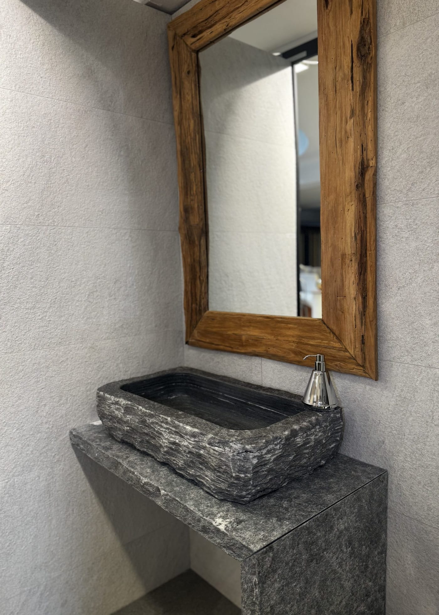 Trog-Waschbecken aus grauem Naturstein auf Steinkonsole, darüber Spiegel mit Altholzrahmen, Wand in großformatigem Feinsteinzeug.