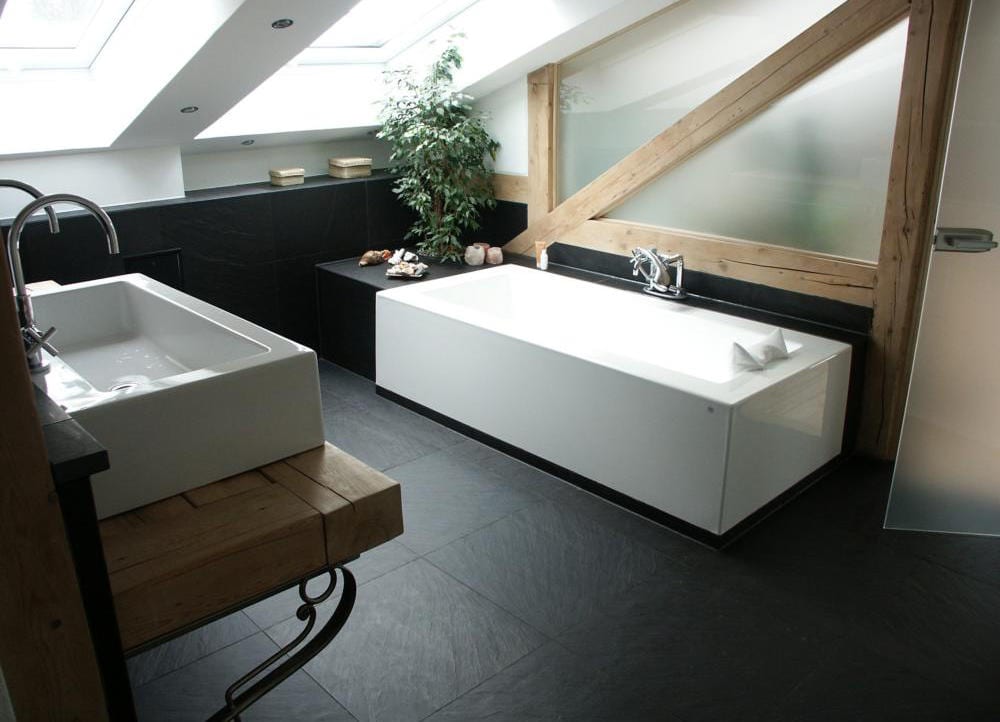 Dachgeschoss-Badezimmer mit anthrazitfarbenem Schieferboden, weißer Wanne und rohen Altholzbalken unter der Dachschräge