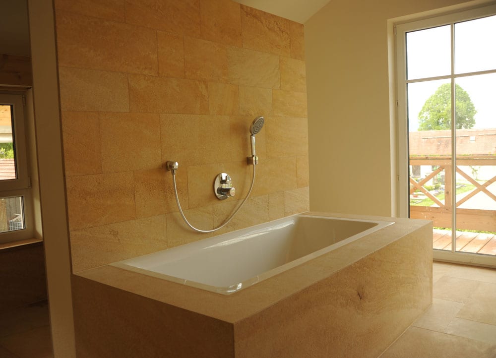 Badewanne mit warm-gelber Sandsteinverkleidung und passenden Wandfliesen, Tageslicht durch bodentiefes Terrassenfenster
