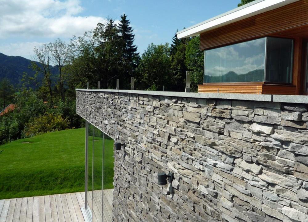 Geschichtete Natursteinmauer an einer Holzterrasse, dahinter Voralpenlandschaft bei Sonnenlicht