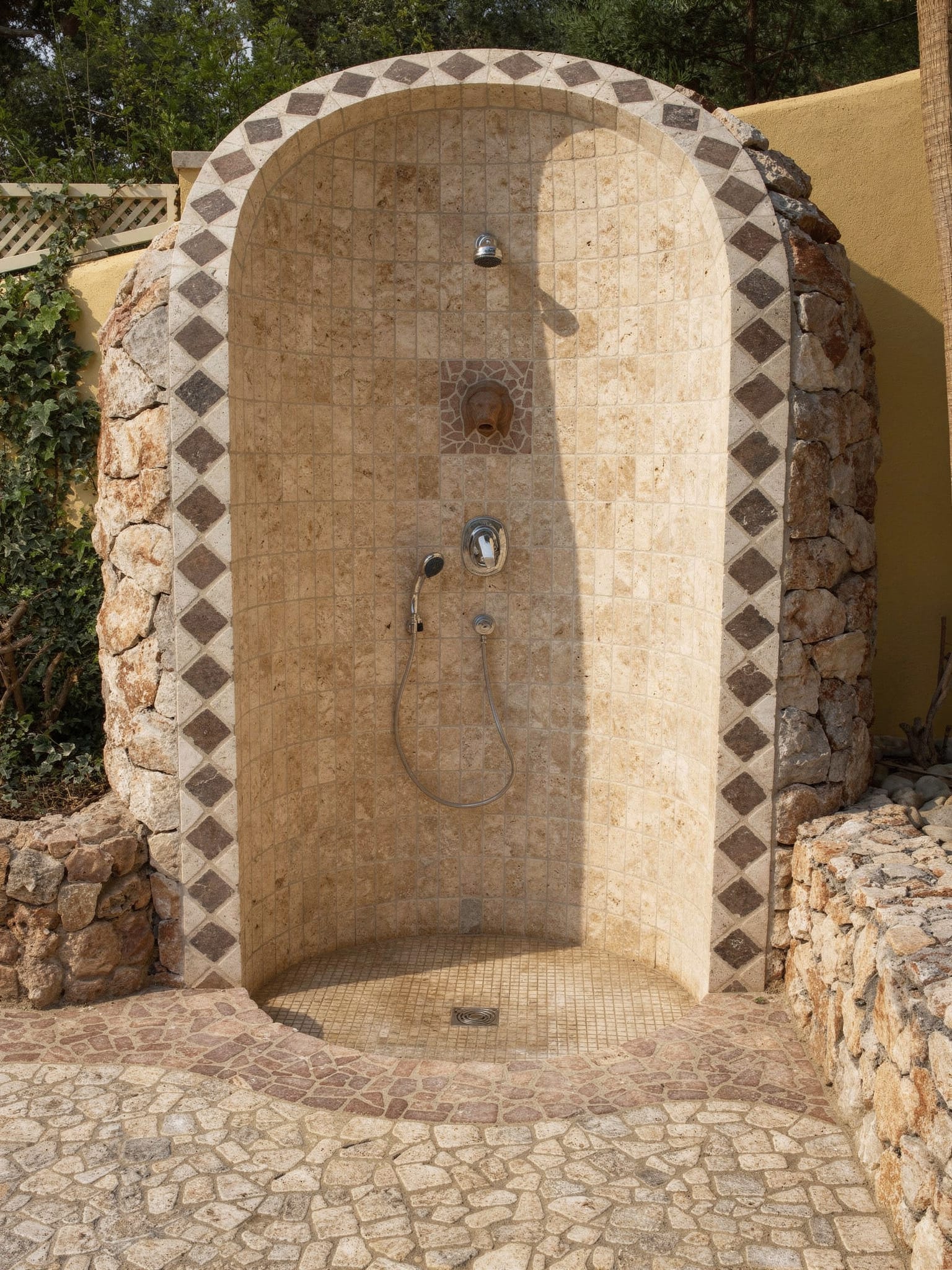 Freistehende Außendusche: gerundete Nische mit Travertin-Mosaik, gerahmt von Natursteinmauerwerk, Bodenmosaik aus Naturstein