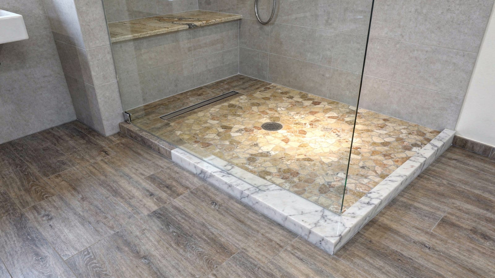 Bodengleiche Dusche mit polygonalem Bruch-Marmormosaik in Beige und Braun, Holzoptik-Dielen davor, Glastrennwand und Sitzbank