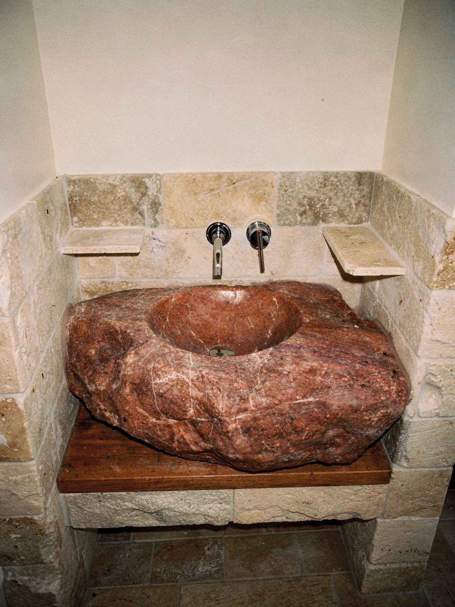 Waschtisch aus einem rotbraun geäderten Naturstein-Findling mit eingearbeitetem Becken, in einer Nische aus hellem Travertin-Mauerwerk