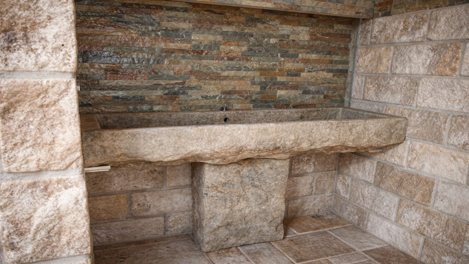 Massives Naturstein-Trogwaschbecken auf Steinsockel vor Schiefer-Mosaikband, umgeben von getrommelten Bruchstein-Wandfliesen