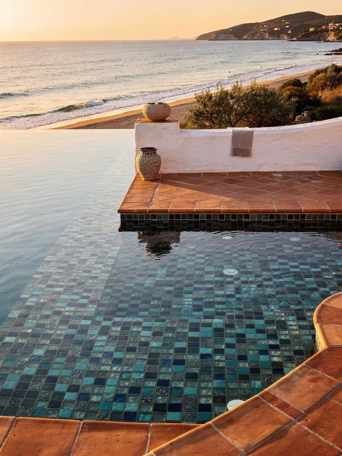 Pool mit blauem Glasmosaik und hellem Natursteinrand vor mediterraner Landschaft